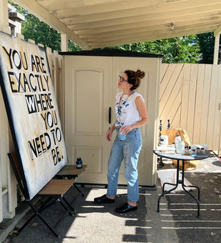 Sara Joy painting in her outdoor studio in New York. In front of her large painting that reads, "You are exactly where you need to be."