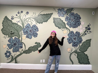 New York Mural Artist, Sara Joy, posing in front of her large botanical mural created for oHHo Botanical Wellness shop in Irvington, New York. The floral mural consists of blue flowers and green leaves.